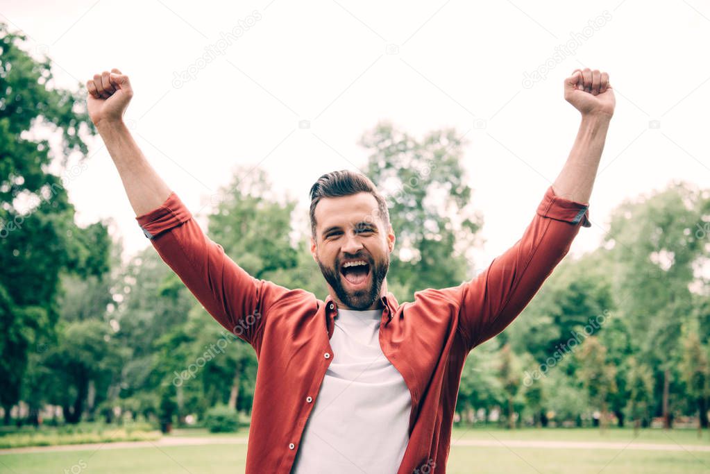 excited young man standing in park and putting hands in air