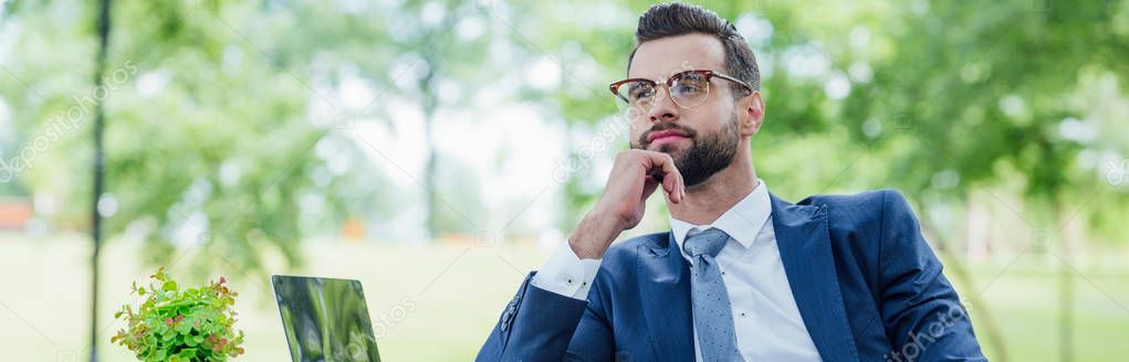 panoramic shot of youg businessman looking away while sitting near table in park