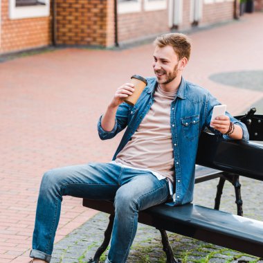 bankta otururken kağıt bardak ve akıllı telefon tutan neşeli adam 