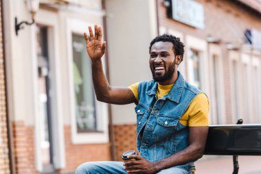 mutlu afrikalı amerikalı adam kağıt bardak tutarken el sallayarak 