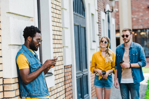 selective focus of handsome african american man using smartphone near friends with paper cups