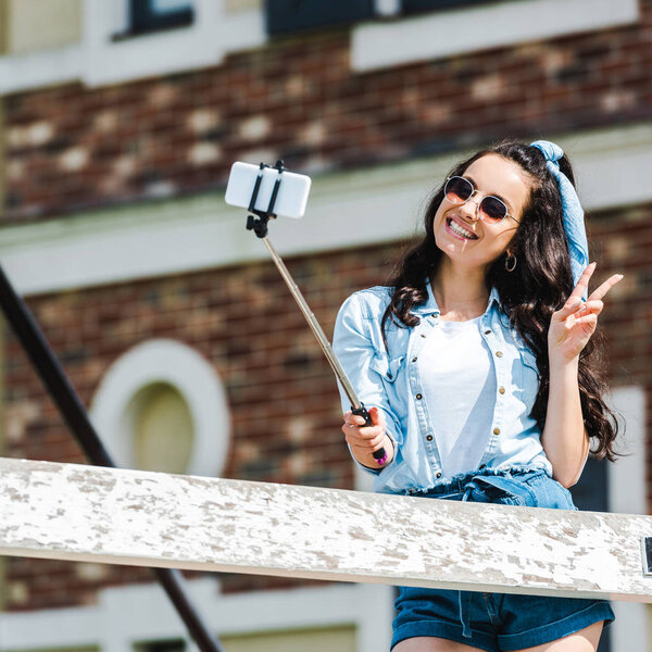 happy woman holding selfie stick and taking selfie while showing peace sign 