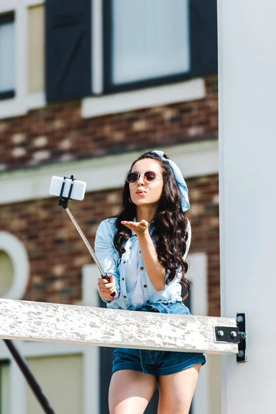 young woman holding selfie stick and taking selfie while sending air kiss