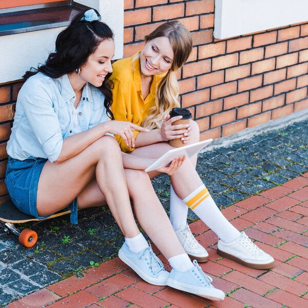 happy woman pointing with finger at digital tablet while sitting on penny board with friend 