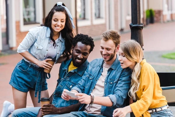 Amigos Multiculturais Felizes Olhando Para Smartphone Enquanto Sentado Banco — Fotografia de Stock
