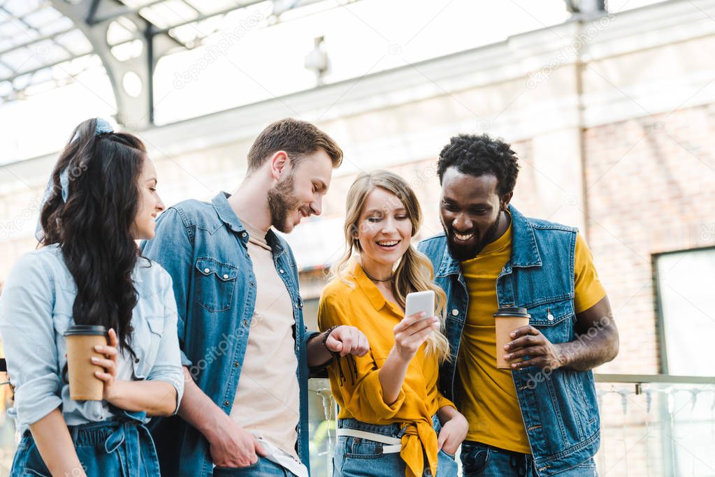 cheerful multicultural friends looking at smartphone while standing together 