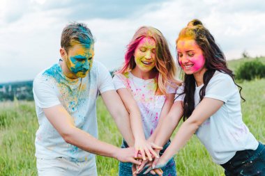 cheerful group of friends with holi paints on faces putting hands together  clipart