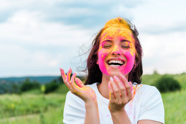happy woman standing with closed eyes and pink and yellow holi paint on face 