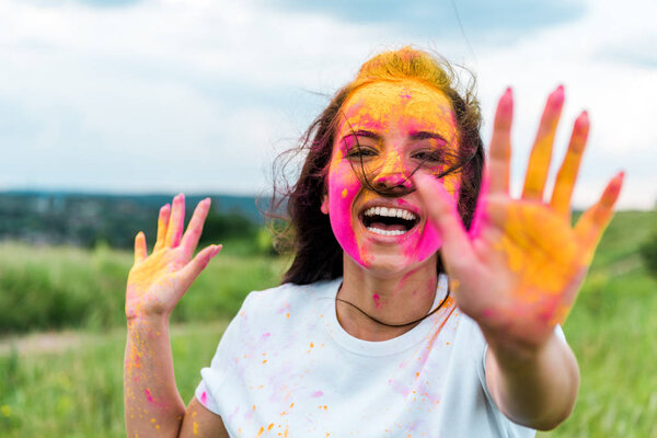 selective focus of happy woman with pink and yellow holi paint on face and hands