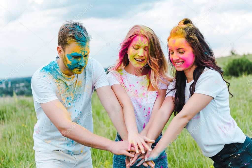 cheerful group of friends with holi paints on faces putting hands together 