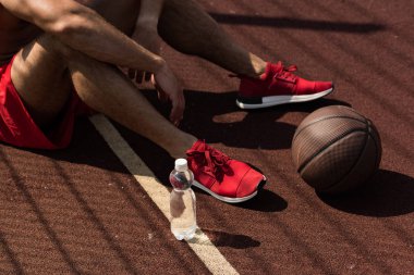 güneşli bir günde su ve top şişesi ile basketbol sahasında oturan kırmızı spor ayakkabılı sporcu kısmi görünümü