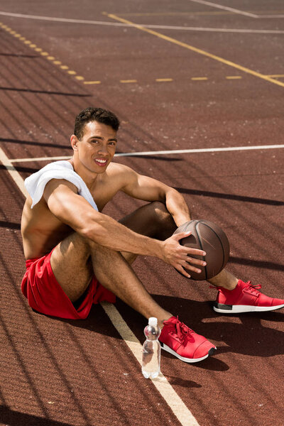 shirtless mixed race basketball player smiling while sitting at basketball court with ball in sunny day