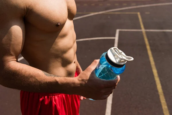 バスケットボールコートでスポーツボトルを保持するシャツなしの筋肉スポーツマンの部分的なビュー — ストック写真
