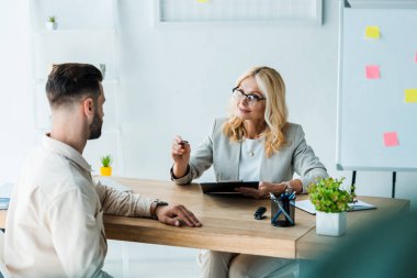 selective focus of blonde recruiter looking at man and holding clipboard with pen  clipart