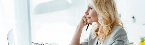 panoramic shot of thoughtful blonde woman holding pen in office 