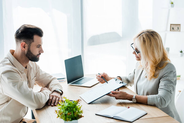 selective focus of cheerful woman holding clipboard and pen near employee and laptop with blank screen 