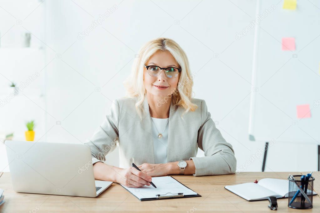 cheerful woman holding pen near clipboard and laptop in office 