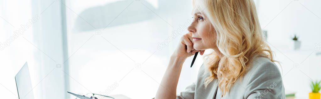 panoramic shot of thoughtful blonde woman holding pen in office 