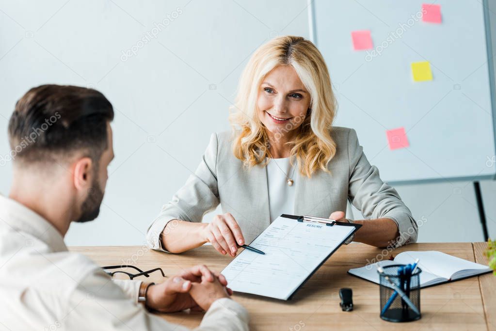 selective focus of happy recruiter holding pen near clipboard and looking at bearded man 