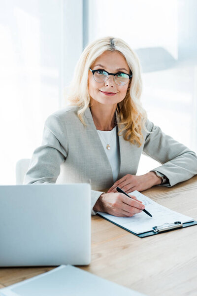 happy blonde woman in glasses looking at camera while holding pen near clipboard and laptop 
