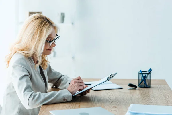 Fröhliche Blonde Frau Mit Brille Blickt Auf Klemmbrett — Stockfoto