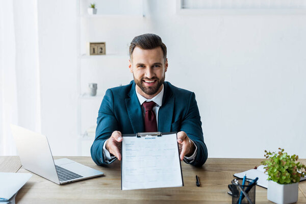 happy recruiter sitting near laptop and holding clipboard with resume