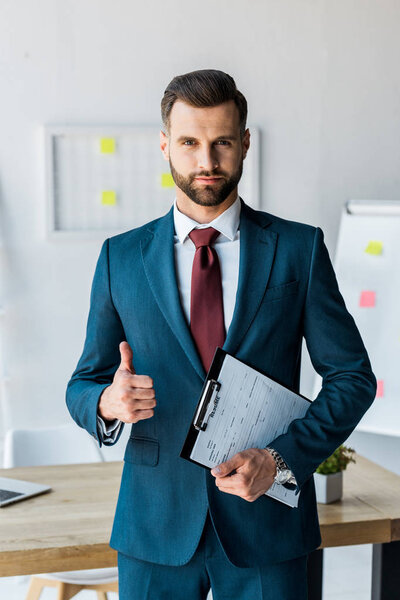 handsome recruiter standing and holding clipboard with resume letters while showing thumb up