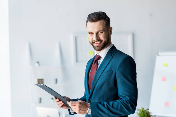 cheerful bearded man in suit holding clipboard in office 