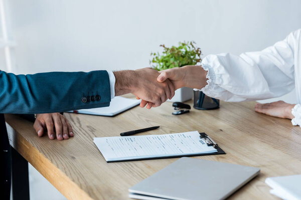 cropped view of recruiter and woman shaking hands near clipboard on table 