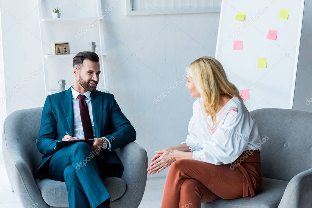 handsome recruiter holding clipboard and looking at blonde employee sitting in armchair 