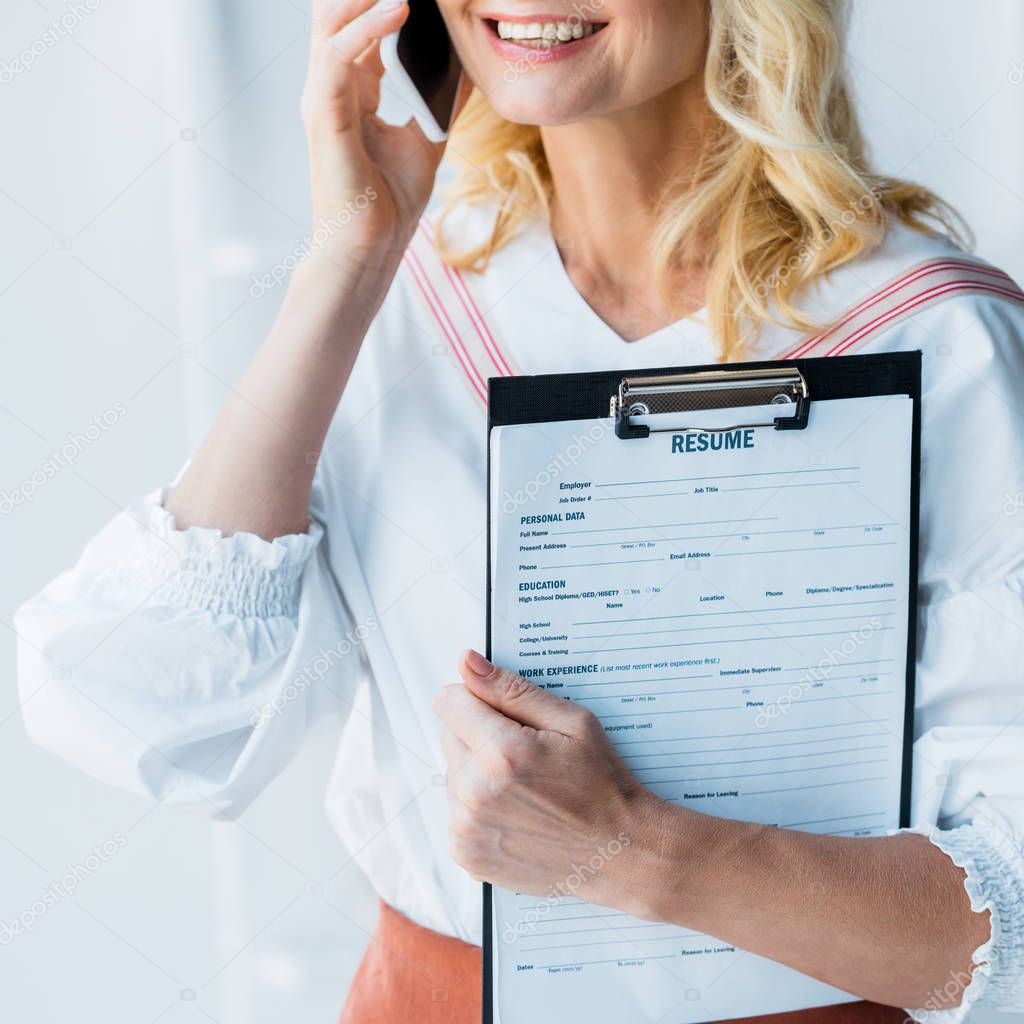 cropped view of happy woman talking on smartphone and holding clipboard with resume letters 