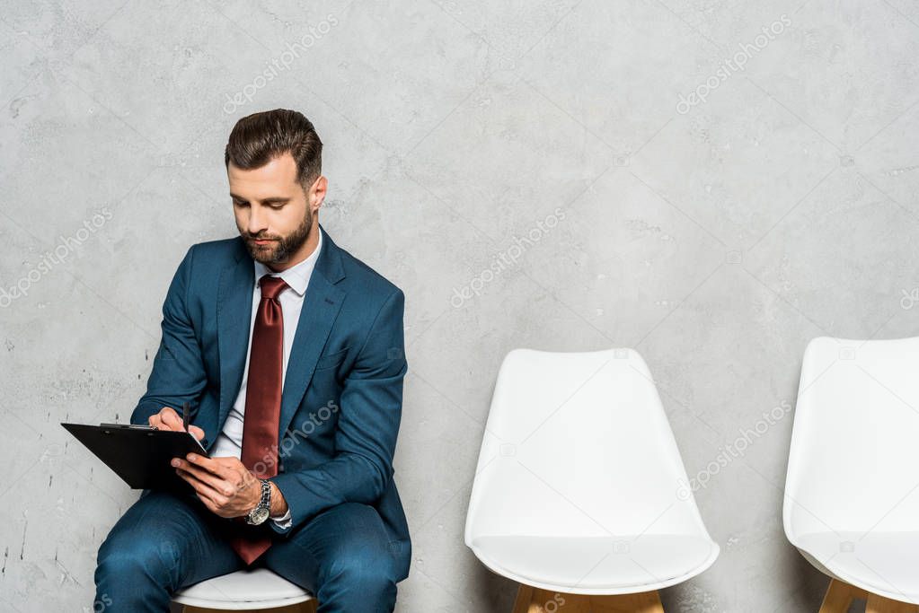 handsome bearded man sitting on white chair and holding clipboard 