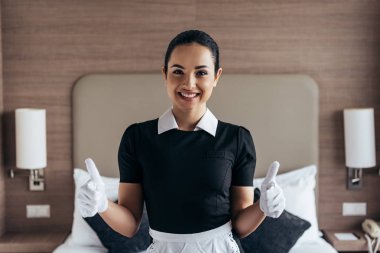 beyaz eldiven ve önlük güzel gülümseyen hizmetçi ön görünümü kamera bakarak ve otel odasında başparmak gösteren