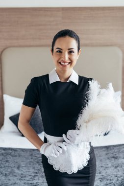 otel odasında yatağın yanında duster tutan beyaz eldiven gülümseyen hizmetçi ön görünümü