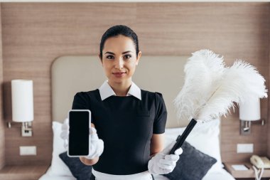 otel odasında boş ekran ile duster ve akıllı telefon tutan beyaz eldiven gülümseyen hizmetçi ön görünümü
