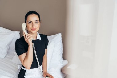 beyaz önlük güzel hizmetçi yatakta oturan ve otel odasında telefonla konuşurken