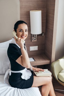 beyaz önlük gülümseyen güzel hizmetçi yatakta oturan ve otel odasında telefonla konuşurken