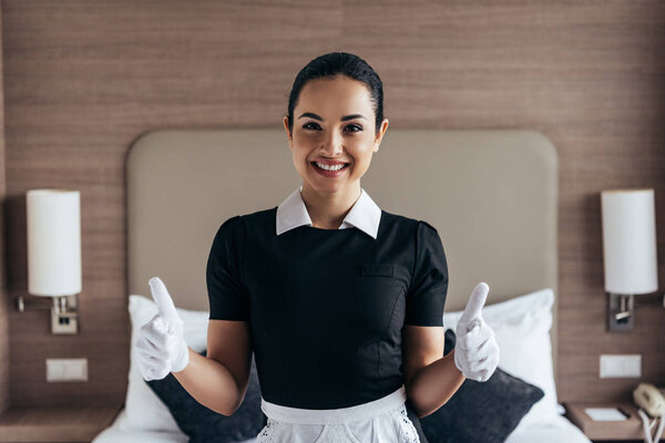 front view of pretty smiling maid in white gloves and apron looking at camera and showing thumbs up in hotel room