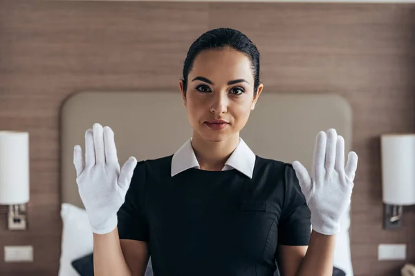 白い手袋とエプロンでカメラを見て ホテルの部屋で手を上げてかわいい笑顔のメイドの正面図 — ストック写真