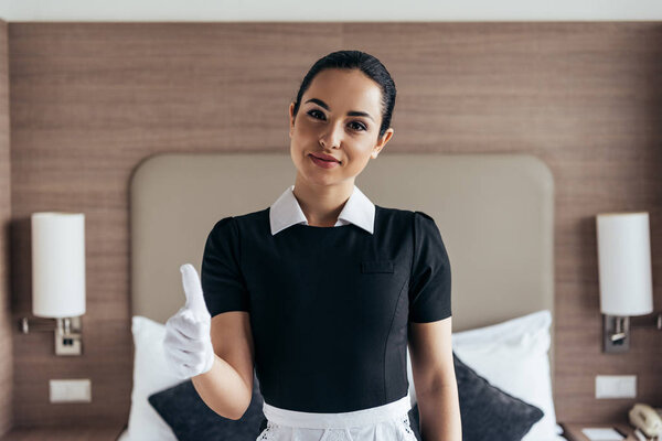 front view of pretty smiling maid in white glove and apron looking at camera and showing thumb up in hotel room