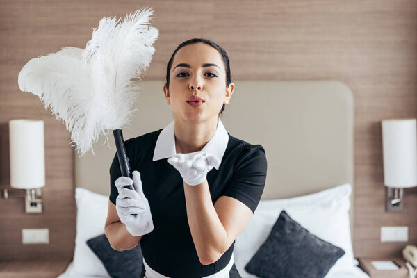 front view of smiling maid in white gloves holding duster and sending air kiss near bed in hotel room