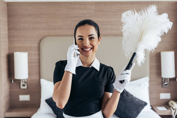 front view of smiling pretty maid in white gloves holding duster and talking on smartphone in hotel room