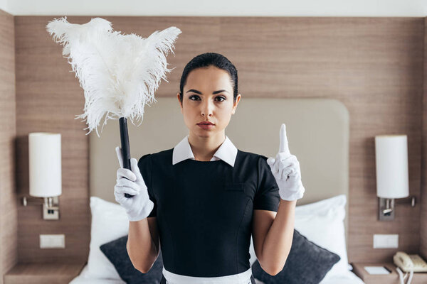 front view of serious maid in white gloves holding duster and showing idea sign in hotel room
