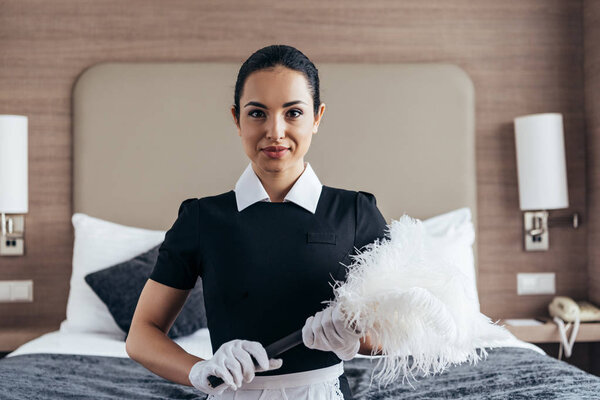 front view of smiling maid in white gloves holding duster near bed and looking at camera