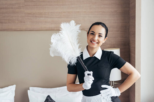 front view of smiling maid in white gloves holding duster near bed and looking at camera