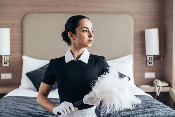 pensive brunette maid in apron and gloves holding duster and looking away in hotel room