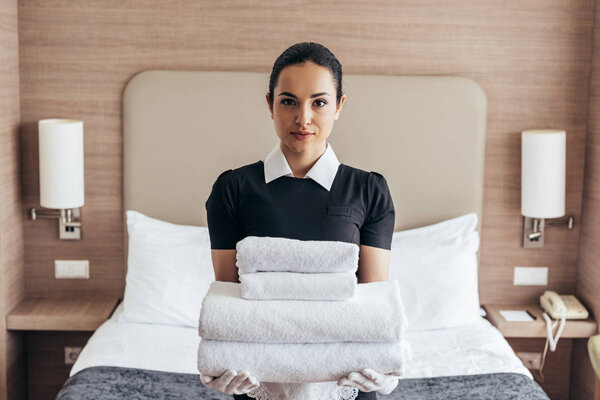 front view of maid holding pile of folded towels near bed and looking at camera in hotel room