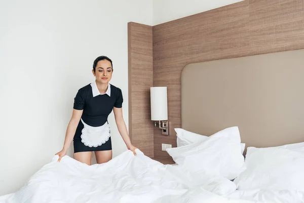 maid in white apron cleaning bed in hotel room