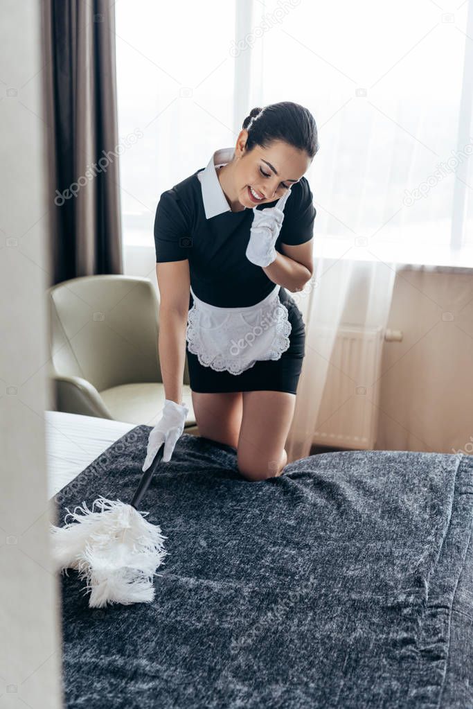 smiling pretty maid in white gloves cleaning bed with duster and talking on smartphone in hotel room