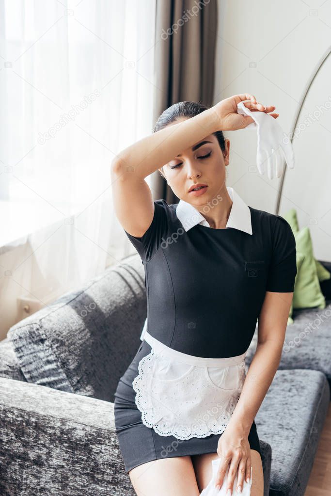 tired maid in white apron sitting on sofa and wiping forehead with closed eyes